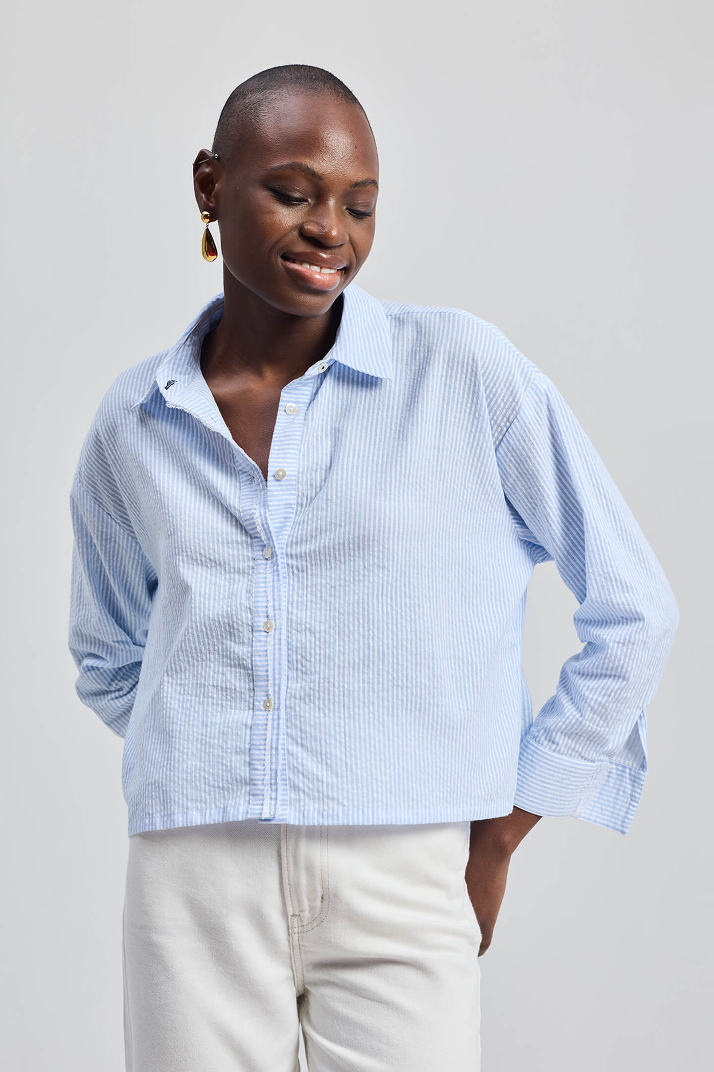 Front view of a smiling model wearing the Reistor Crop it like it’s Hot Shirt in Stripes, featuring blue and white vertical stripes, a button-down front, and a relaxed cropped fit paired with white trousers.