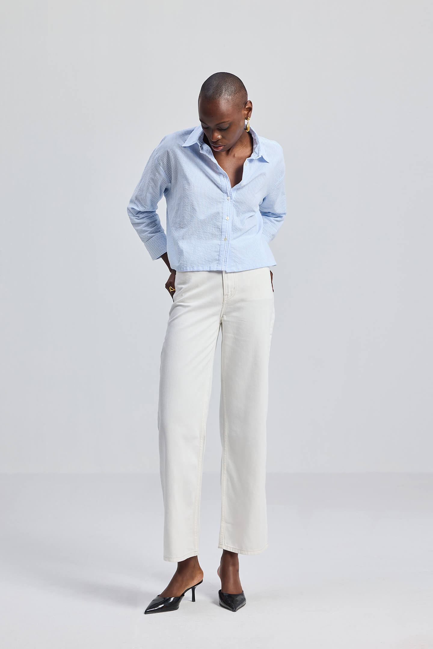 A model wearing the Reistor Crop it like it’s Hot Shirt in Stripes, featuring light blue and white vertical stripes, a relaxed cropped fit, and a classic button-down front paired with white trousers.