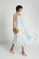 Woman wearing the Reistor Dancing in the Moonlight Dress, a light blue floral midi dress, holding a straw clutch against a white background for effortless summer style.