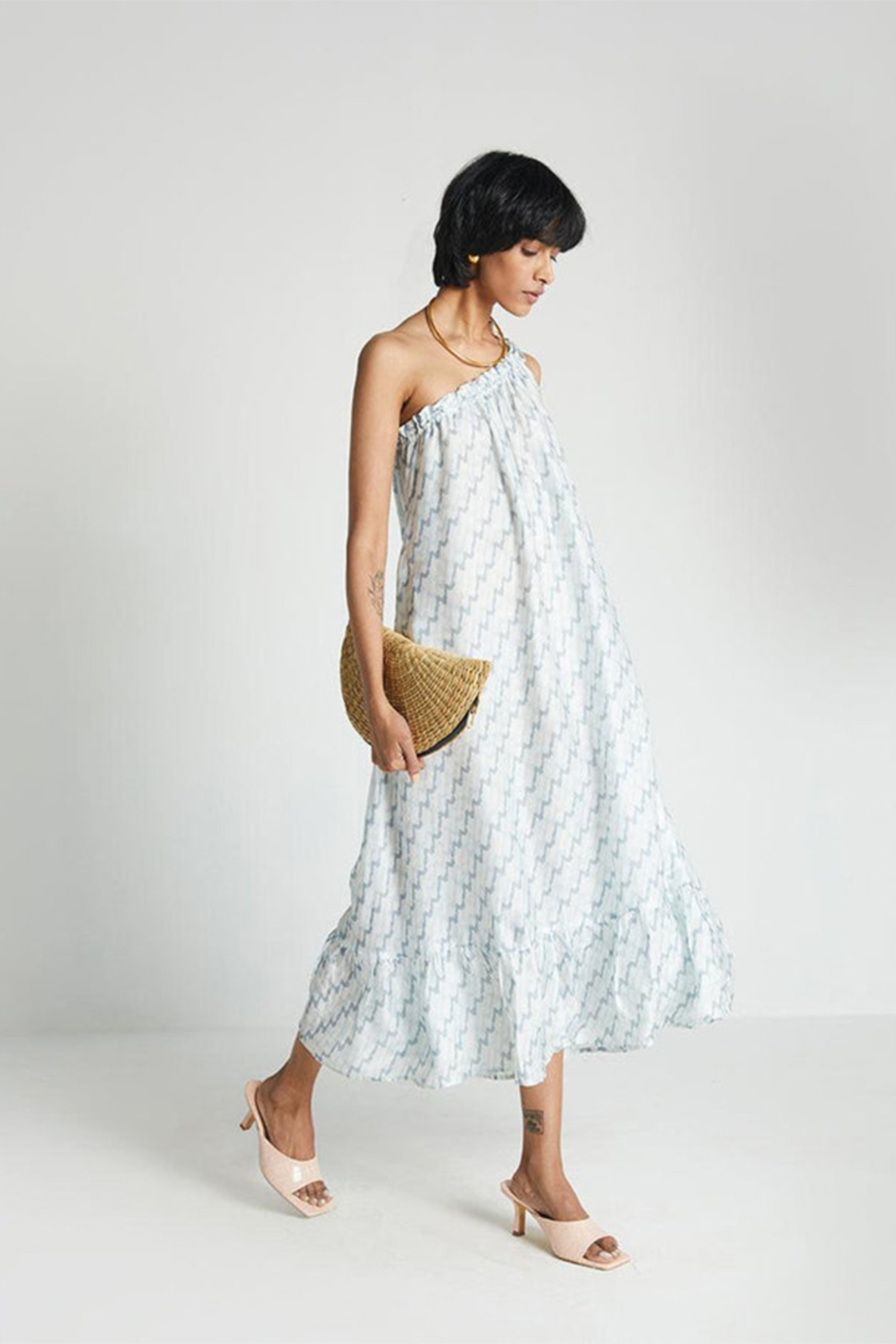 Woman wearing the Reistor Dancing in the Moonlight Dress, a light blue floral midi dress, holding a straw clutch against a white background, perfect for summer outings.