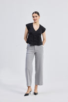 Deep V-neck short top in Black by reistor featuring cap sleeves, front button detailing, and a relaxed fit silhouette styled with grey wide-leg trousers, full-length view.