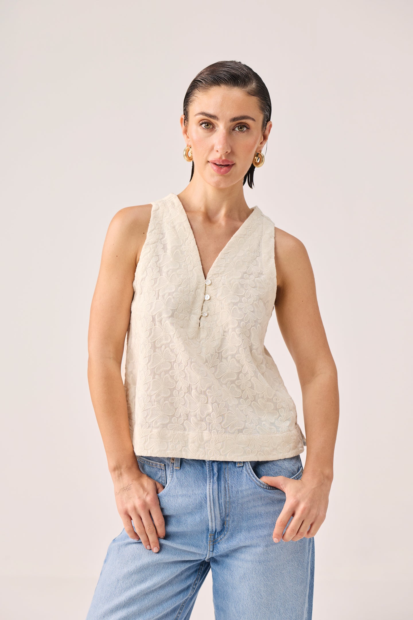 Embroidered Button Front Neck Top in Cream featuring intricate floral embroidery, a sleeveless silhouette, and a V-neckline with pearl-style button detailing, shown from the front and styled with light-wash denim jeans in a minimalist studio setting. 