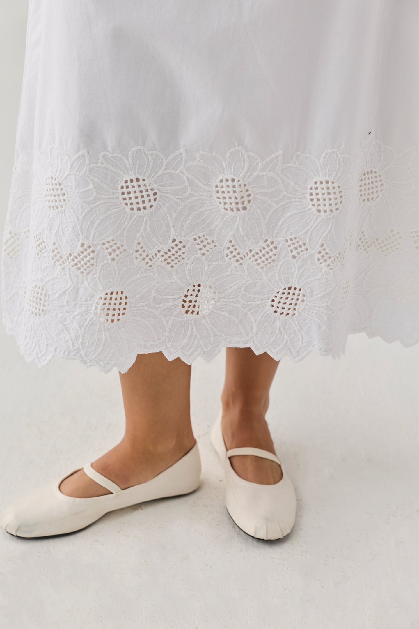 Embroidered Midi Skirt in White featuring a floral eyelet lace hem with scalloped detailing, shown worn with white Mary Jane flats. The skirt falls to mid-calf length and has a light, airy silhouette.