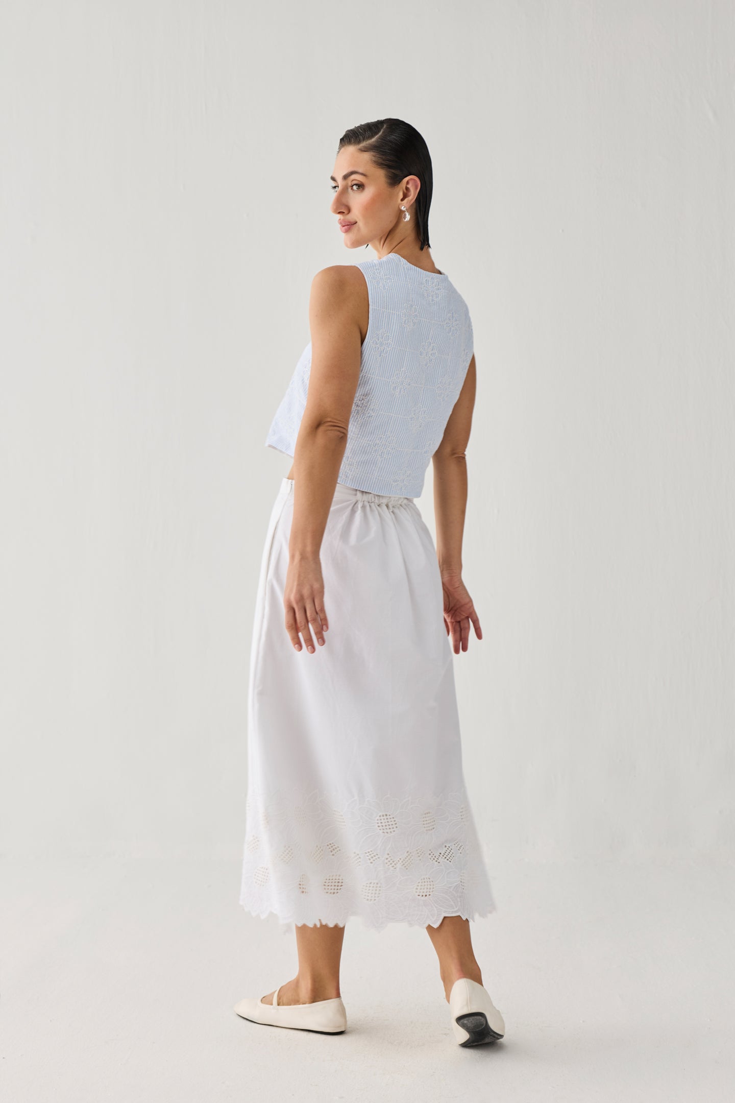 Embroidered Midi Skirt in White — White midi skirt with delicate embroidered eyelet detailing along the hem, styled with a light blue sleeveless textured top and white flats, shown from the back in a minimalist studio setting.