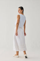 Embroidered Midi Skirt in White — White midi skirt with delicate embroidered eyelet detailing along the hem, styled with a light blue sleeveless textured top and white flats, shown from the back in a minimalist studio setting.
