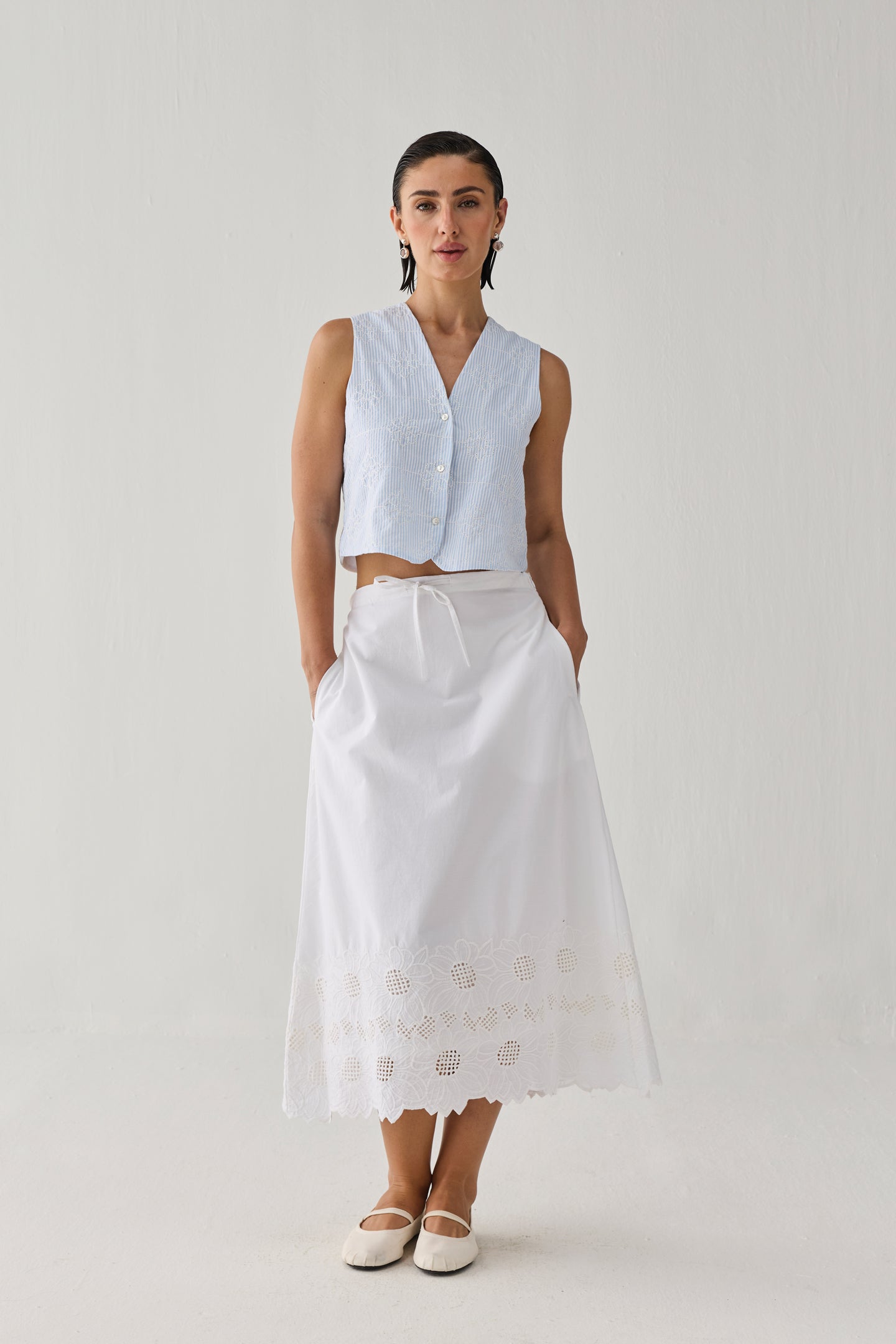 Woman wearing an embroidered white midi skirt with delicate cutwork detailing along the hem, paired with a sleeveless light blue striped top and white flats, standing against a plain light background.