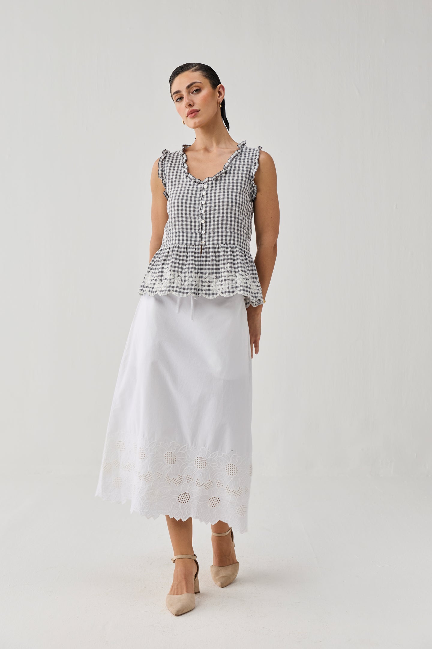 Woman wearing an embroidered peplum top in a grey check pattern with ruffled edges and front button detailing, paired with a white midi skirt featuring embroidered cutwork at the hem and beige pointed-toe heels, standing against a plain light background.