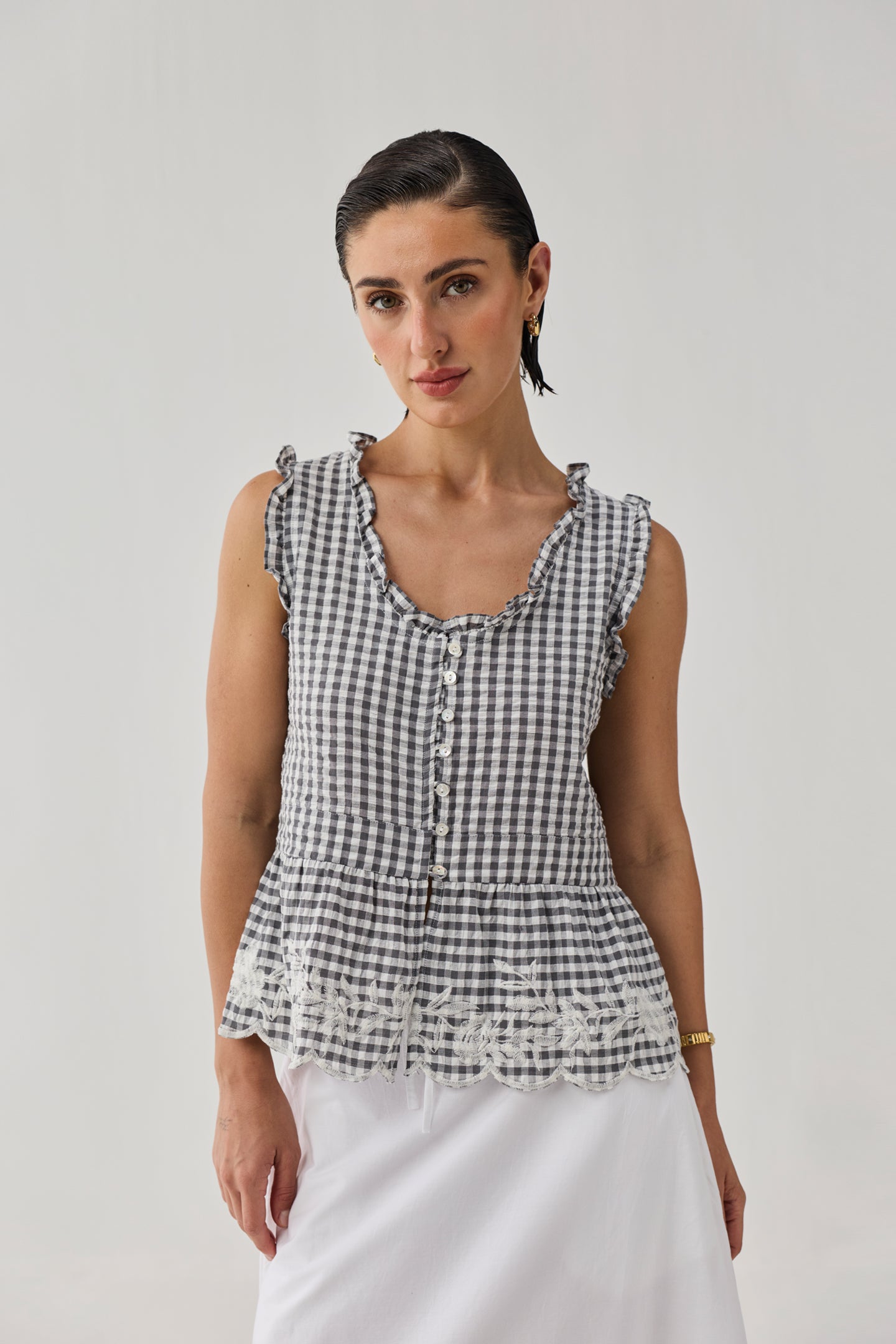 Woman wearing an embroidered peplum top in grey gingham check with ruffled trim, front button detailing, and lace embroidery at the hem, paired with a white skirt against a neutral background.