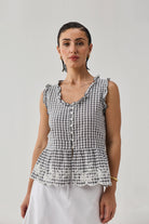 Model wearing an embroidered peplum top in grey check, featuring a gingham pattern, ruffled neckline and armholes, front button closure, and delicate lace embroidery along the peplum hem, paired with a white skirt.