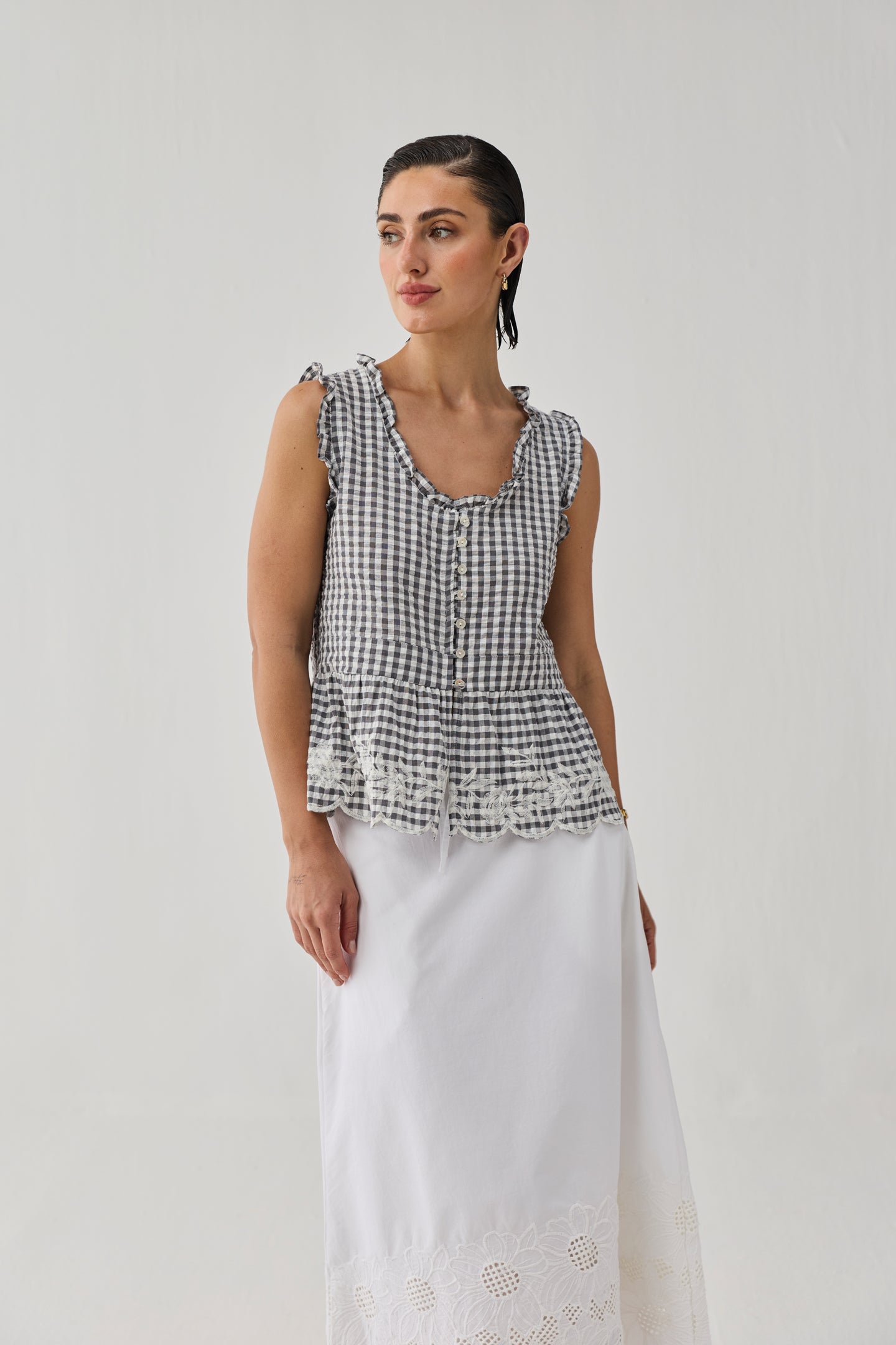 Full-length view of a model wearing an embroidered peplum top in grey check, featuring a gingham pattern, ruffled edges, front button detailing, and lace embroidery along the hem. The top is paired with a flowing white skirt and photographed against a clean, neutral backdrop.