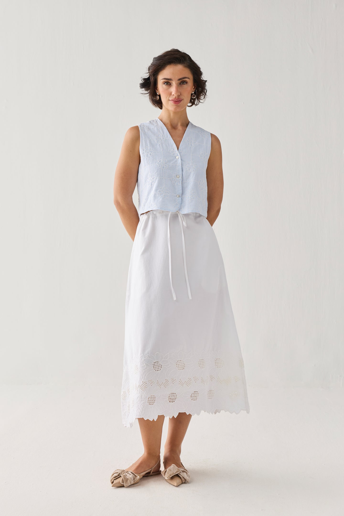Embroidered Sleeveless Vest Top in Blue Stripe featuring delicate tonal embroidery, a subtle textured stripe pattern, V-neckline, and button-front closure, styled with a white embroidered midi skirt and photographed in a full-length studio portrait.