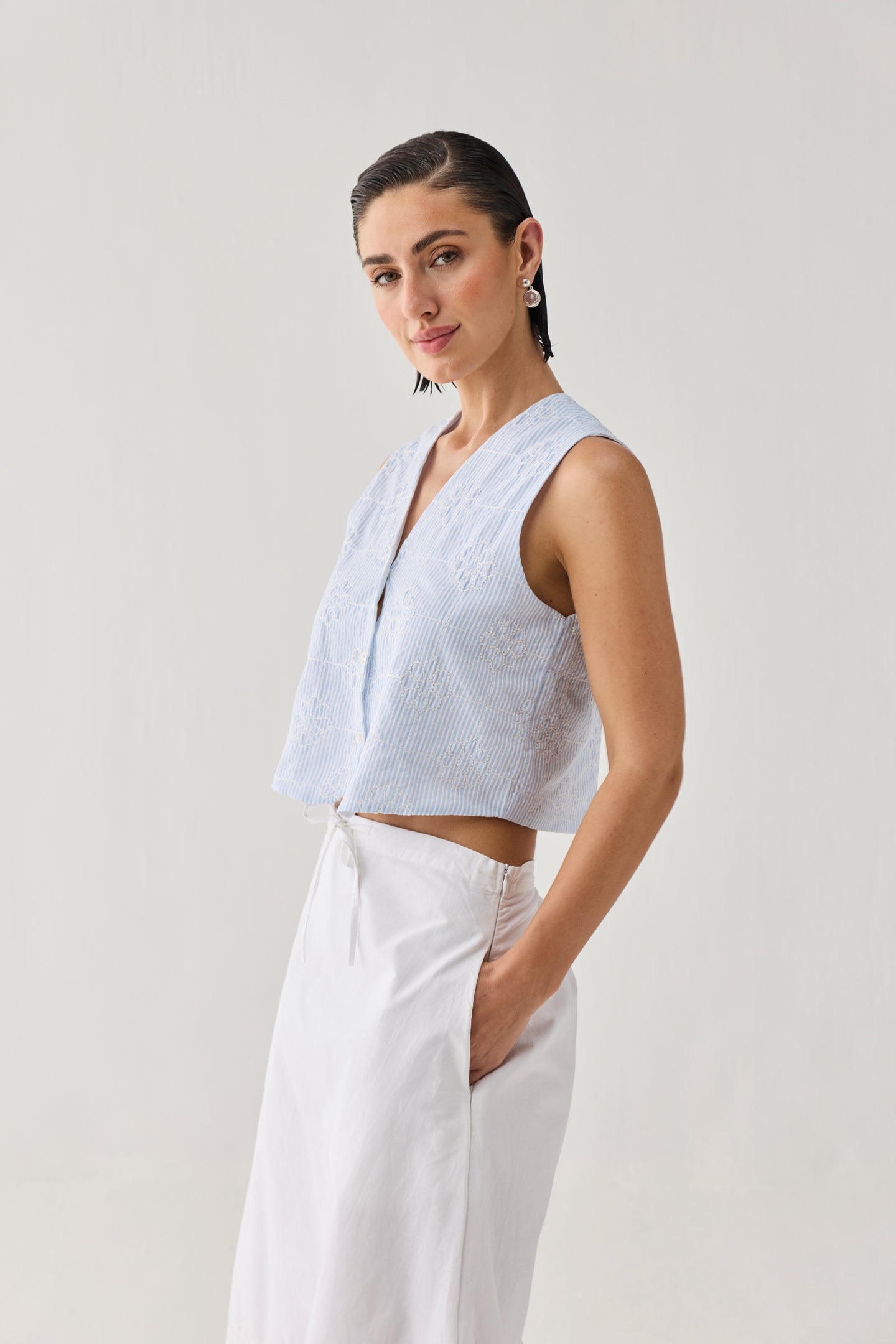 Embroidered Sleeveless Vest Top in Blue Stripe with a textured striped pattern, V-neckline, and cropped sleeveless silhouette, shown from a side angle and paired with a white skirt in a minimalist studio setting.