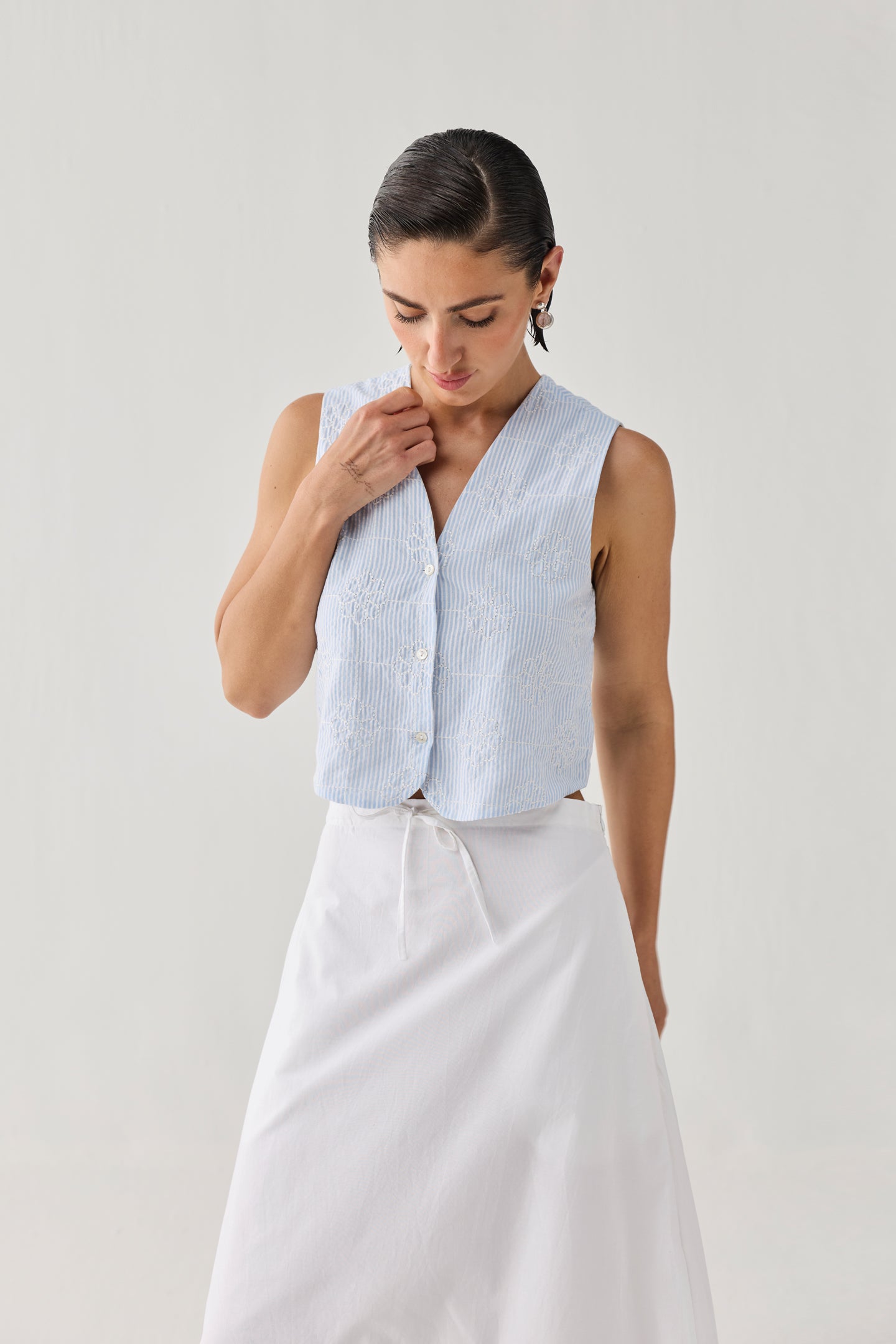 Embroidered Sleeveless Vest Top in Blue Stripe featuring a subtle textured stripe pattern, V-neckline, button-front closure, and sleeveless cropped fit, styled with a white skirt and photographed in a clean studio setting.