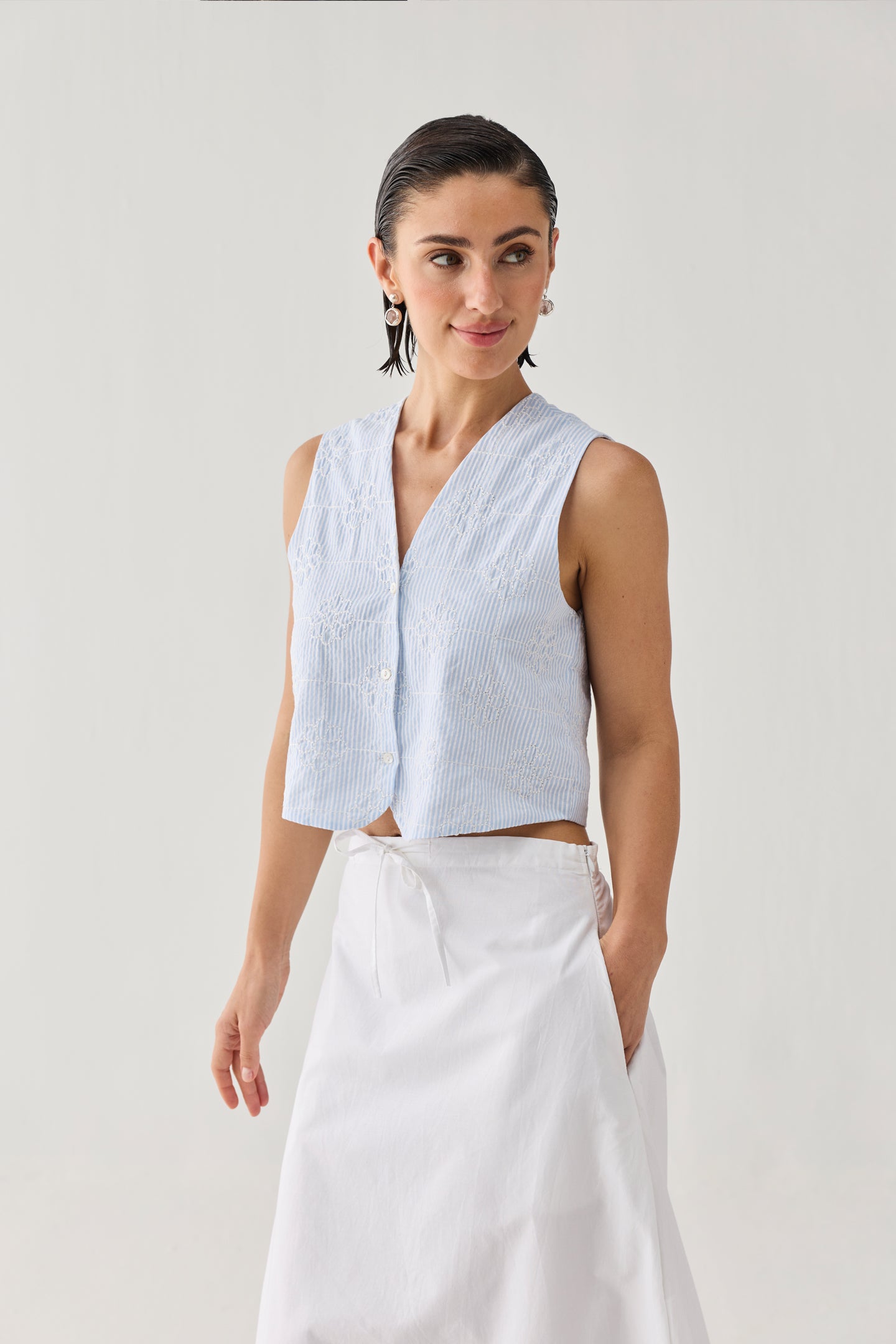 Embroidered Sleeveless Vest Top in Blue Stripe featuring a subtle textured stripe pattern, delicate tonal embroidery, a V-neckline, and button-front closure, styled with a white skirt and shown in a full-length studio portrait.