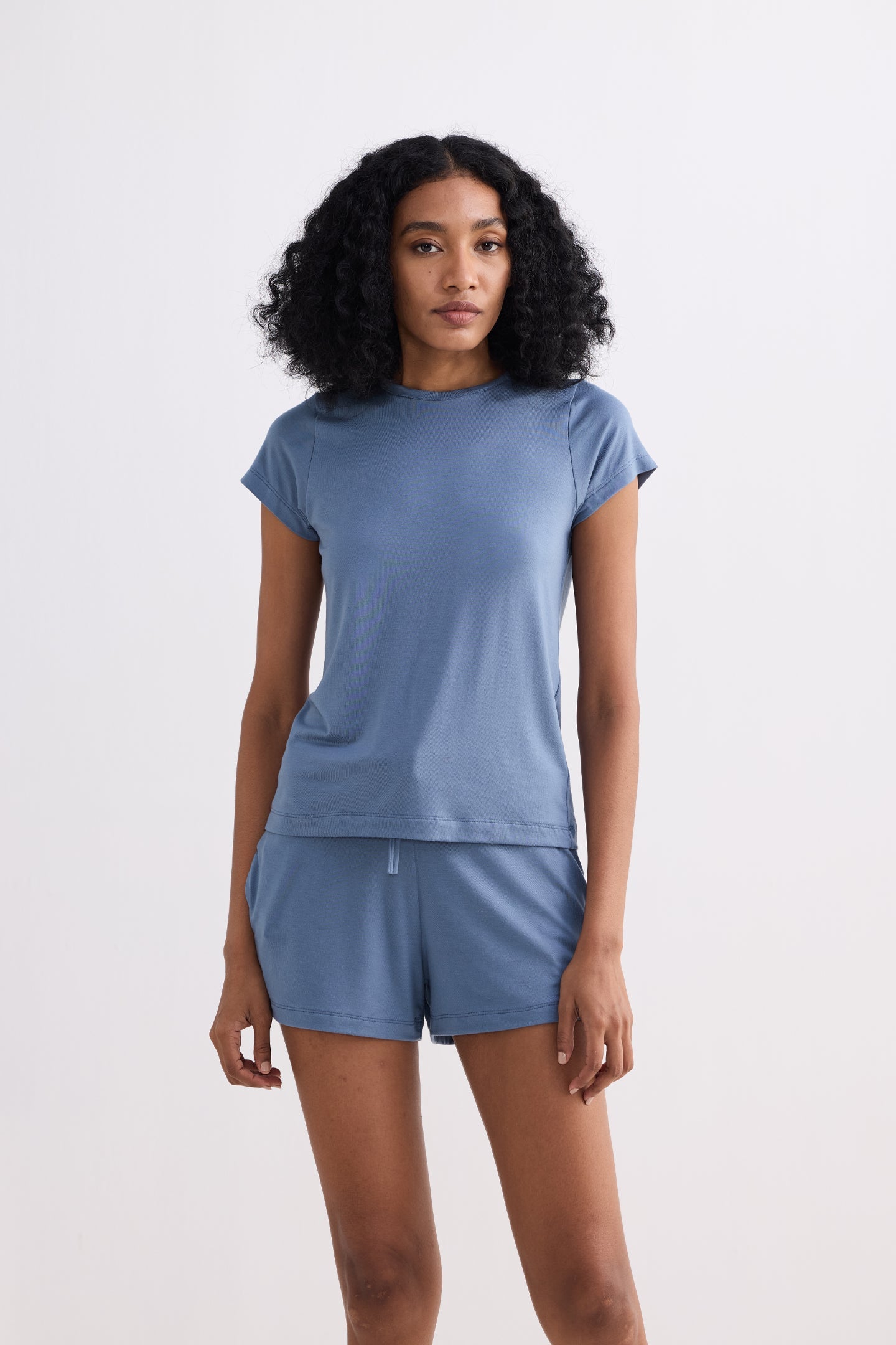 A full-body studio view of a model in the reistor Essential Short Sleeve Tee Set in Blue, featuring a slim-profile crew neck top and coordinating mini-shorts crafted from soft, sustainable blue jersey fabric.