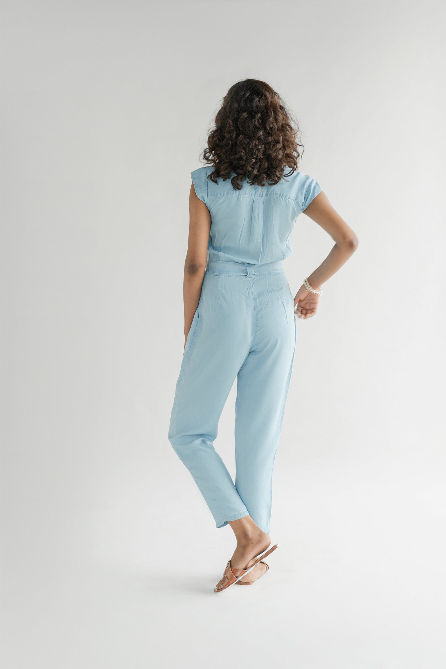 Back view of Reistor Evening Chai Jumpsuit in Blue Denim featuring a relaxed fit, cap sleeves, tapered legs, and a breathable sustainable design for everyday wear.
