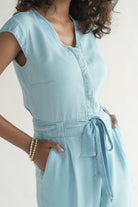 Close-up of Reistor Evening Chai Jumpsuit in Blue Denim featuring a V-neck, cap sleeves, adjustable tie waist, side pockets, and soft sustainable fabric.