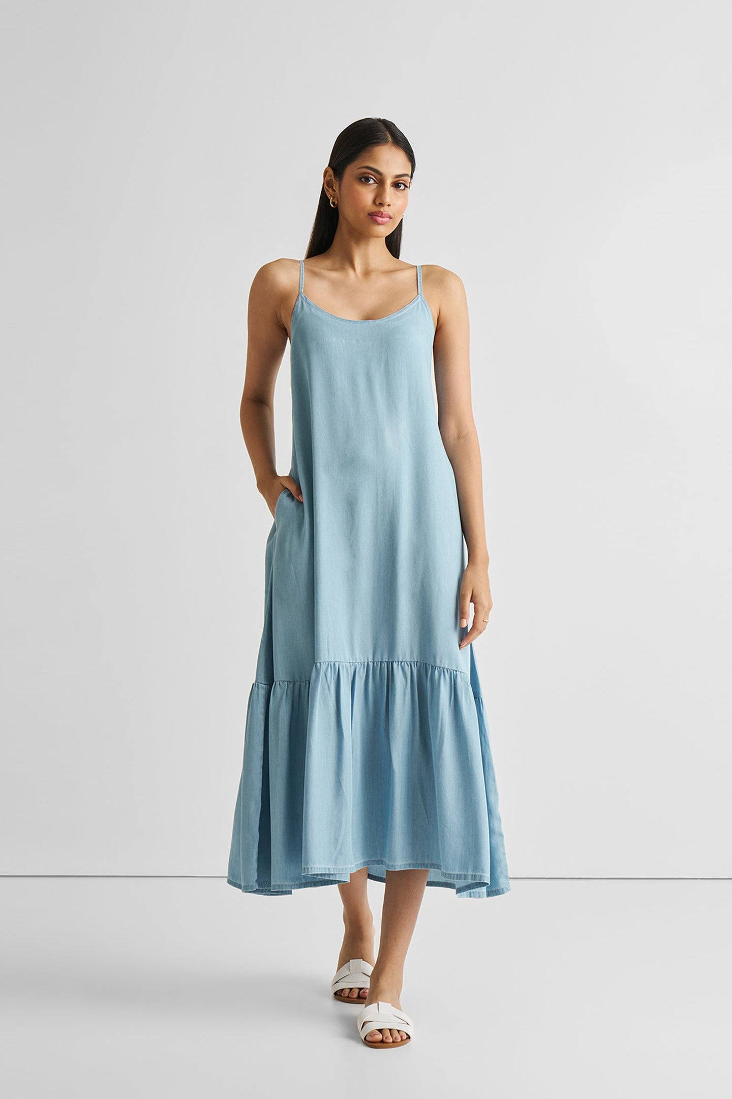 A woman standing and modeling a blue denim maxi dress with a flowy design and adjustable straps, paired with white sandals.