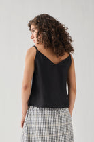 Hemp Front Tie Cami Top in Black, shown from the back, featuring a relaxed fit, thin shoulder straps, and a lightweight breathable fabric. The model pairs it with a check-pattern skirt.