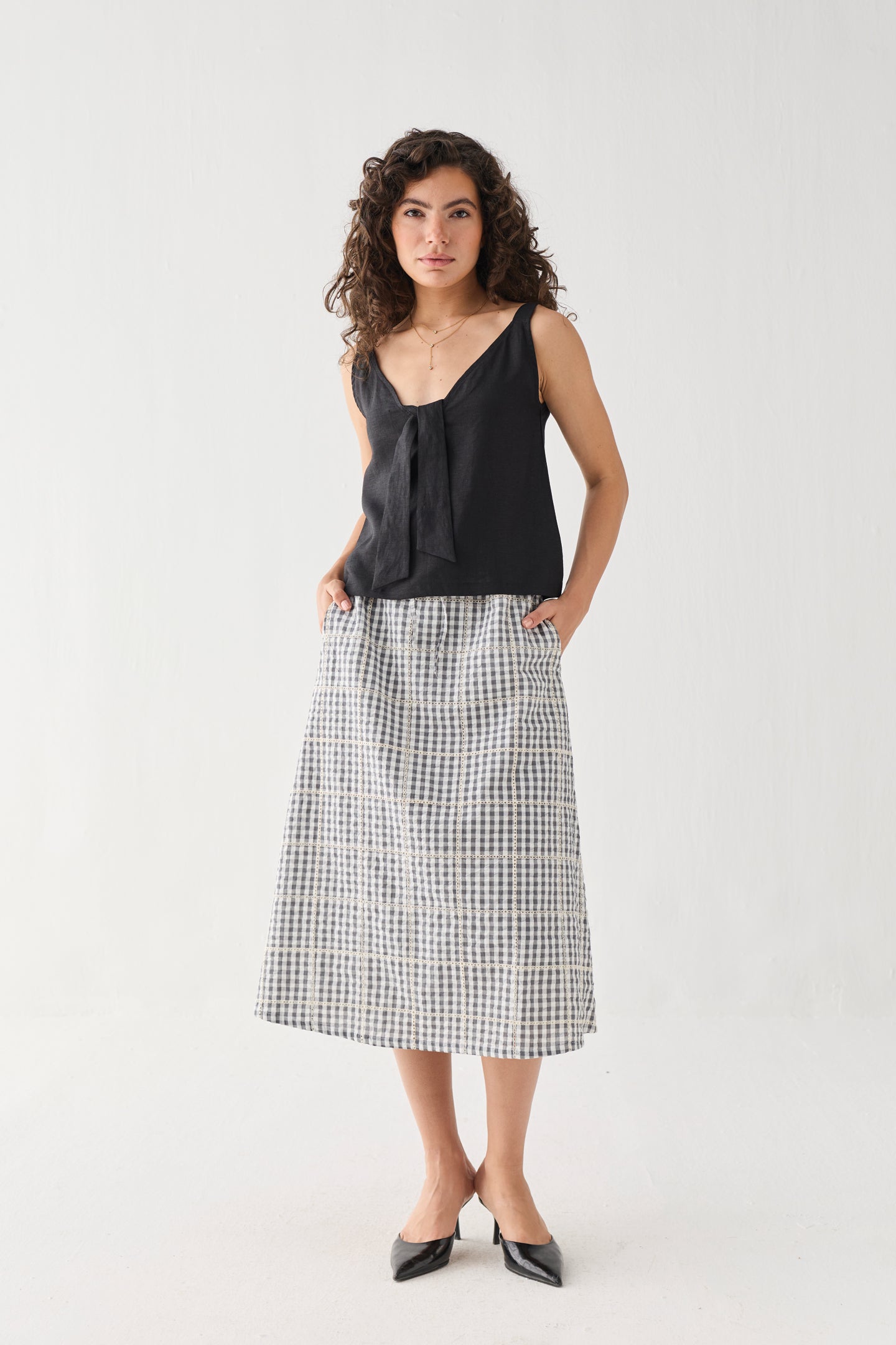 Full-length front view of the Hemp Front Tie Cami Top in Black, featuring a relaxed silhouette, sleeveless design, and elegant front tie detail at the neckline. Made from breathable hemp fabric, the top is styled with a check-pattern midi skirt and pointed flats.
