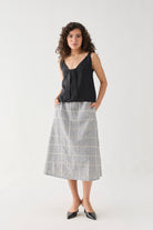 Full-length front view of the Hemp Front Tie Cami Top in Black, featuring a relaxed silhouette, sleeveless design, and elegant front tie detail at the neckline. Made from breathable hemp fabric, the top is styled with a check-pattern midi skirt and pointed flats.