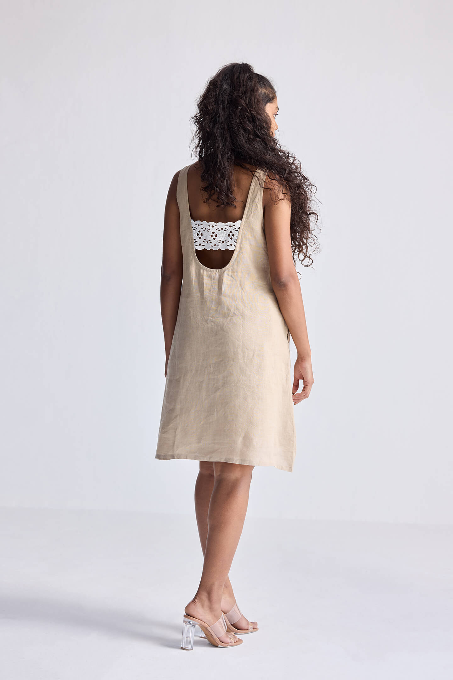 Model wearing the reistor Hemp Short Dress with Back Embroidered Lace Detail, featuring a natural beige hemp fabric, sleeveless silhouette, open back design, and delicate embroidered lace, styled with minimal heels.