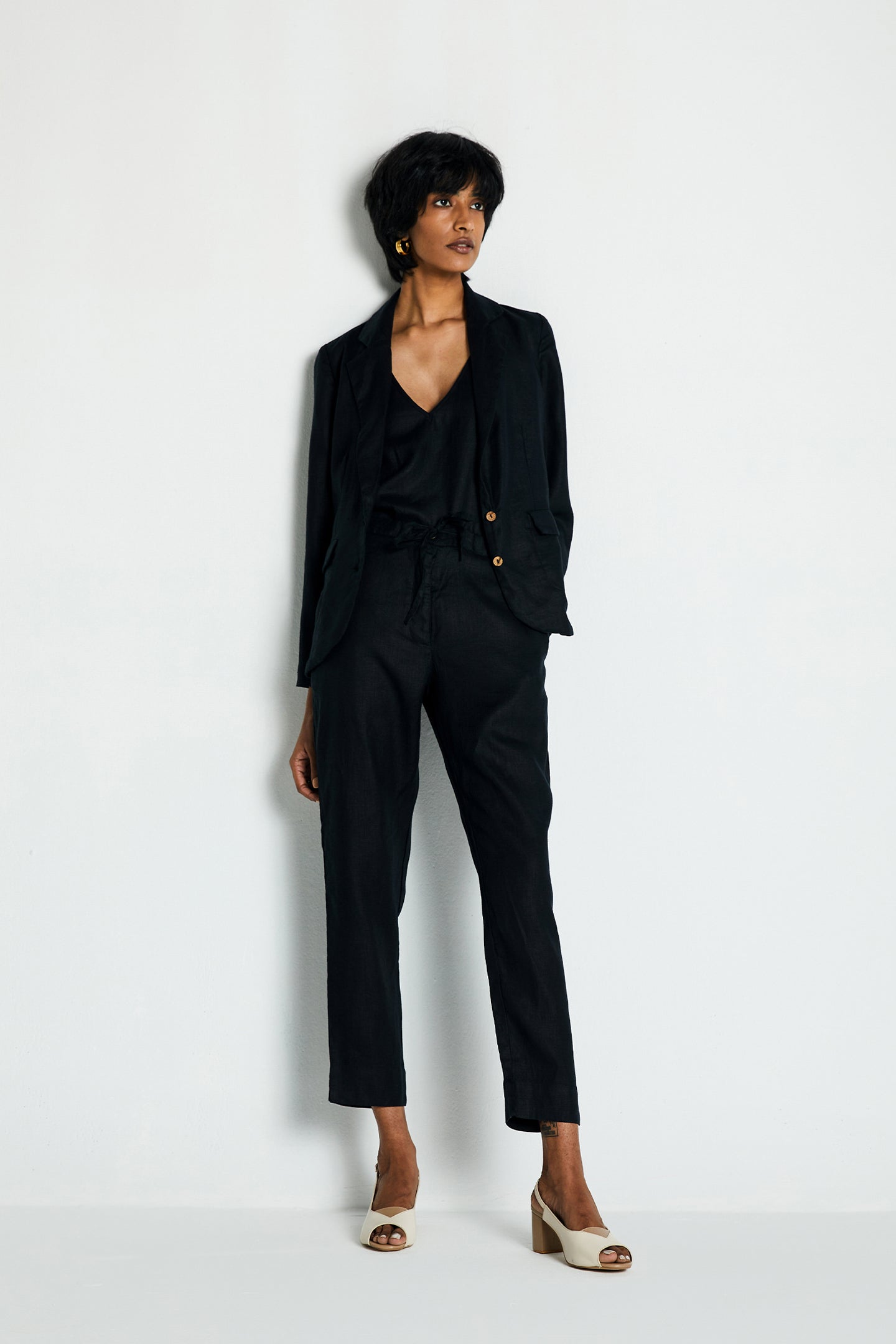 A woman standing against a white background, wearing a black three piece hemp suit consisting of a blazer, top, and pants.