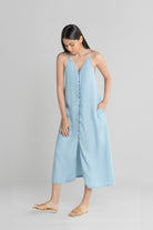 A woman standing and facing to the side, wearing a blue denim maxi dress with a V-neckline, sleeveless design, and ankle length. She has a tan belt and is wearing sandals.