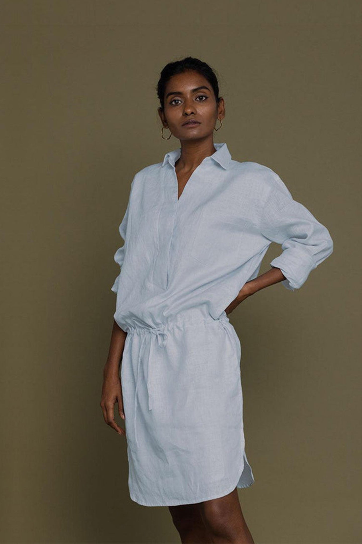 A woman standing against a plain background, wearing a blue collared shirt dress with a front pocket and drawstring waist.
