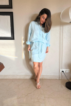 Woman wearing a light blue outfit standing against a neutral wall.