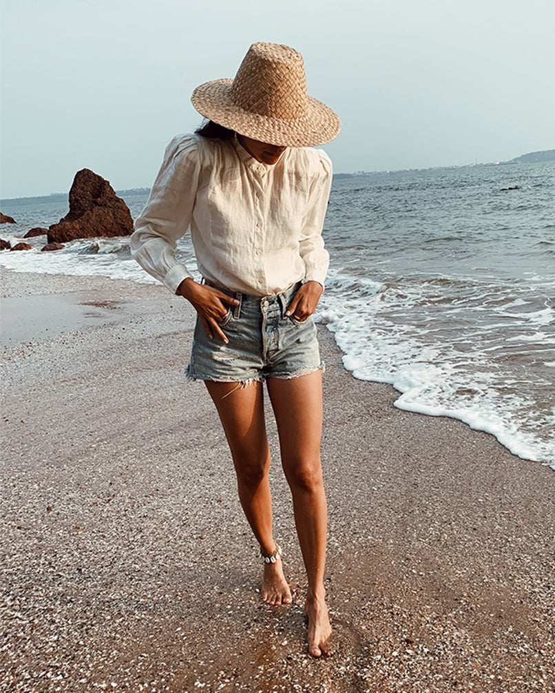 Person wearing a straw hat and denim shorts standing on a beach with ocean waves.
