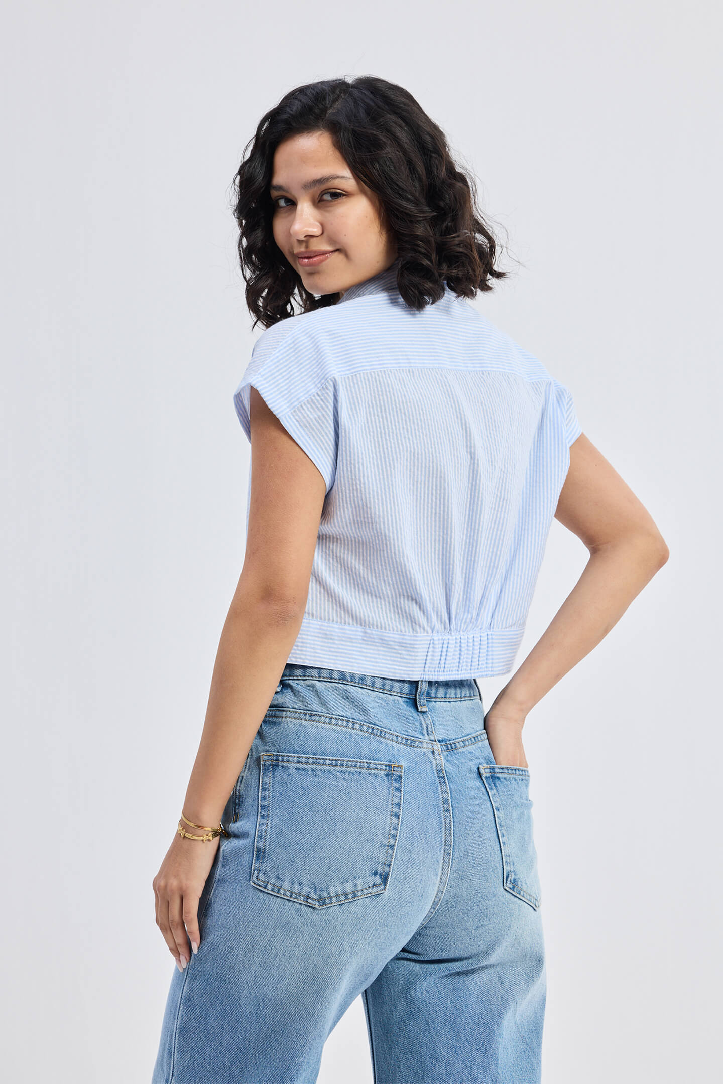 Back view of a model wearing the Reistor Oversized Crop Shirt in Classic Stripes, a light blue and white striped short-sleeve button-down with an elasticated back hem, styled with blue jeans.