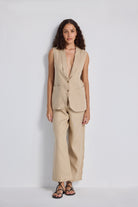 Front view of a model with hands on her hips wearing the beige Sleeveless Hemp Jacket Set by reistor, featuring a button-up longline vest and matching wide-leg trousers.