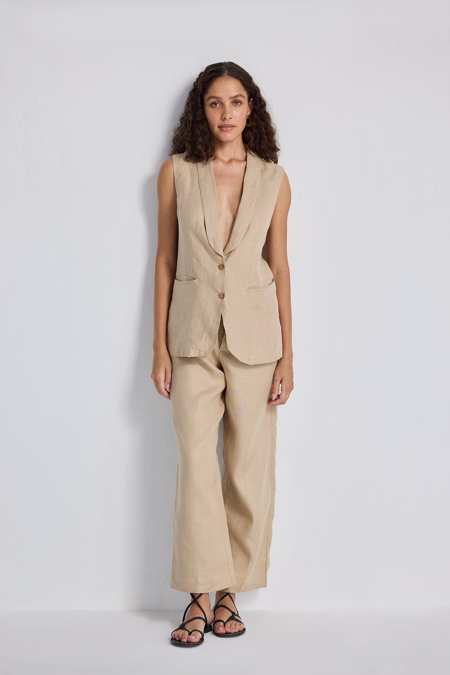 Front view of a model with hands on her hips wearing the beige Sleeveless Hemp Jacket Set by reistor, featuring a button-up longline vest and matching wide-leg trousers.