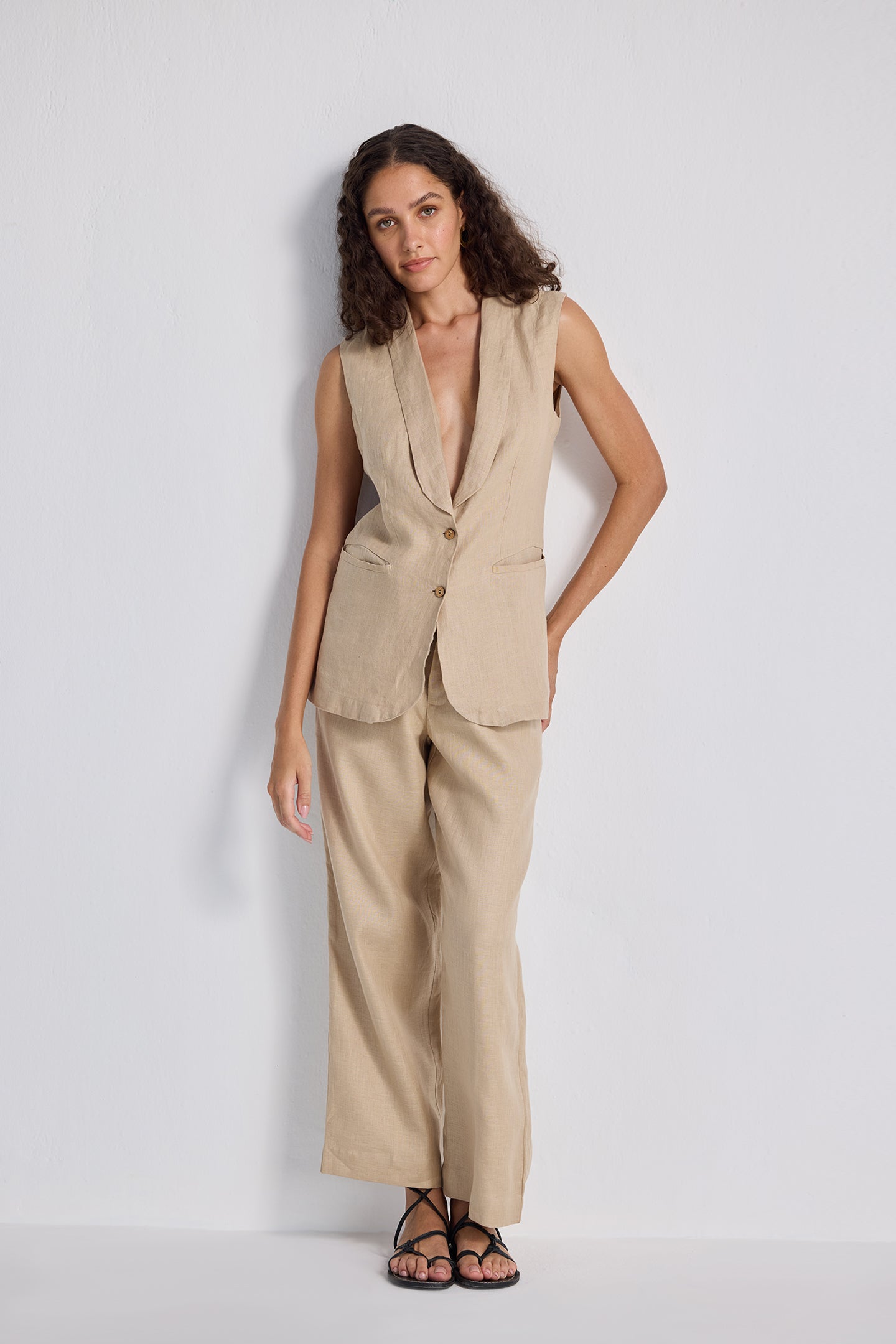 Model wearing the Sleeveless Hemp Jacket Set by Reistor in a natural beige tone, featuring a longline tailored vest with a notched lapel and matching wide-leg trousers, styled with black strappy sandals.