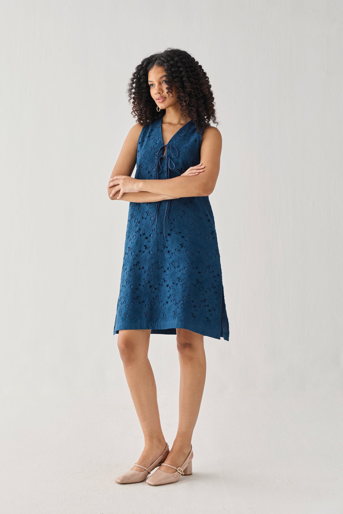 Sleeveless Knee-Length Dress in Blue featuring intricate all-over eyelet embroidery, a V-neckline with self-tie detail, and a relaxed A-line silhouette, shown in a full-length studio portrait that highlights its effortless fit and elegant texture.