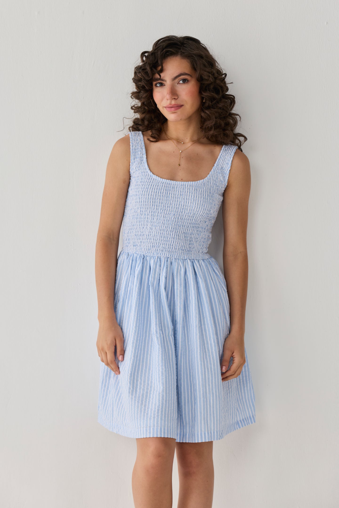 Smocked Short Dress in Blue Stripes designed with a fitted textured bodice and airy striped skirt, creating a timeless casual-chic outfit. 