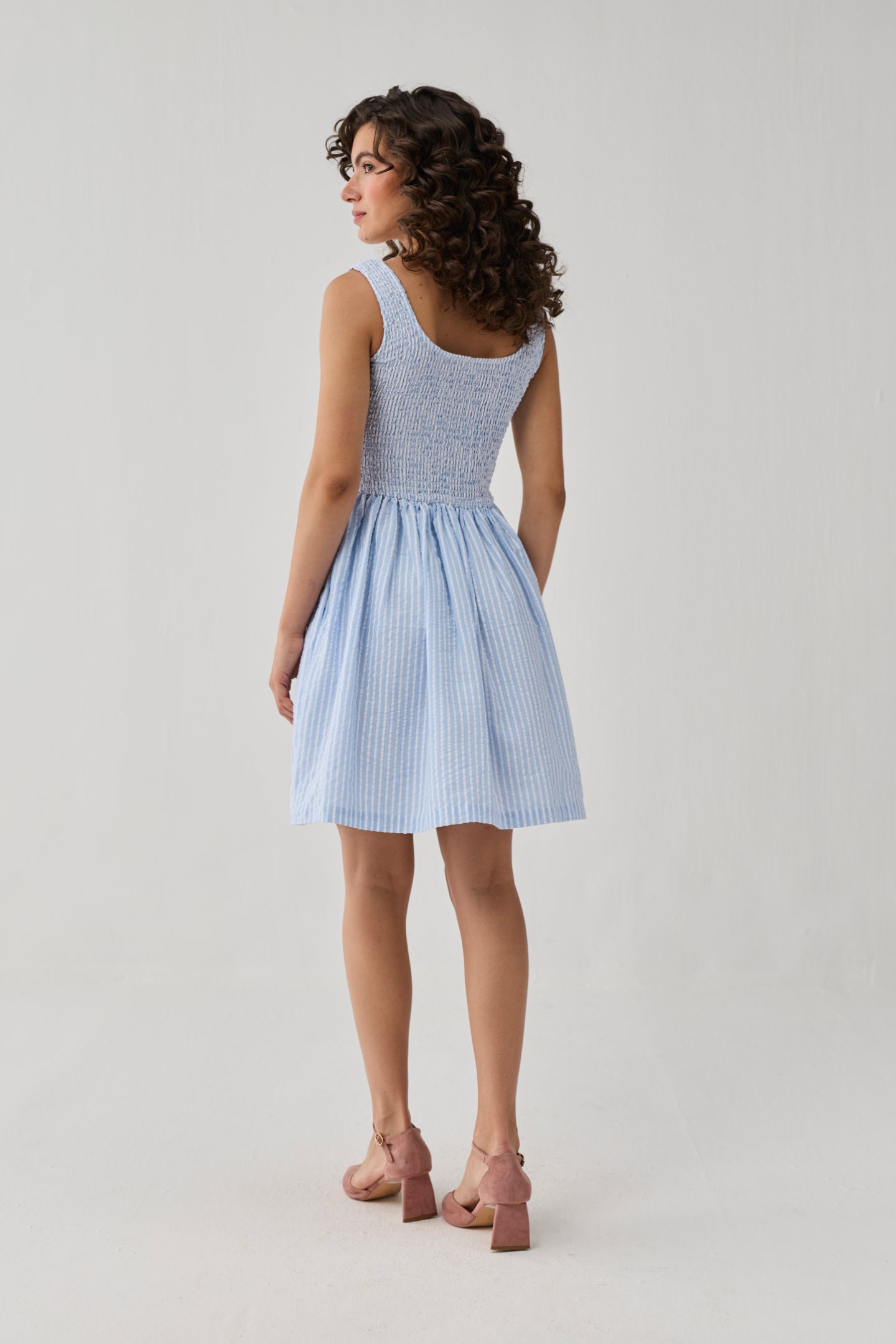 Back view of a light blue striped short dress with a fully smocked bodice, wide straps, and a softly flared skirt for an airy summer feel.