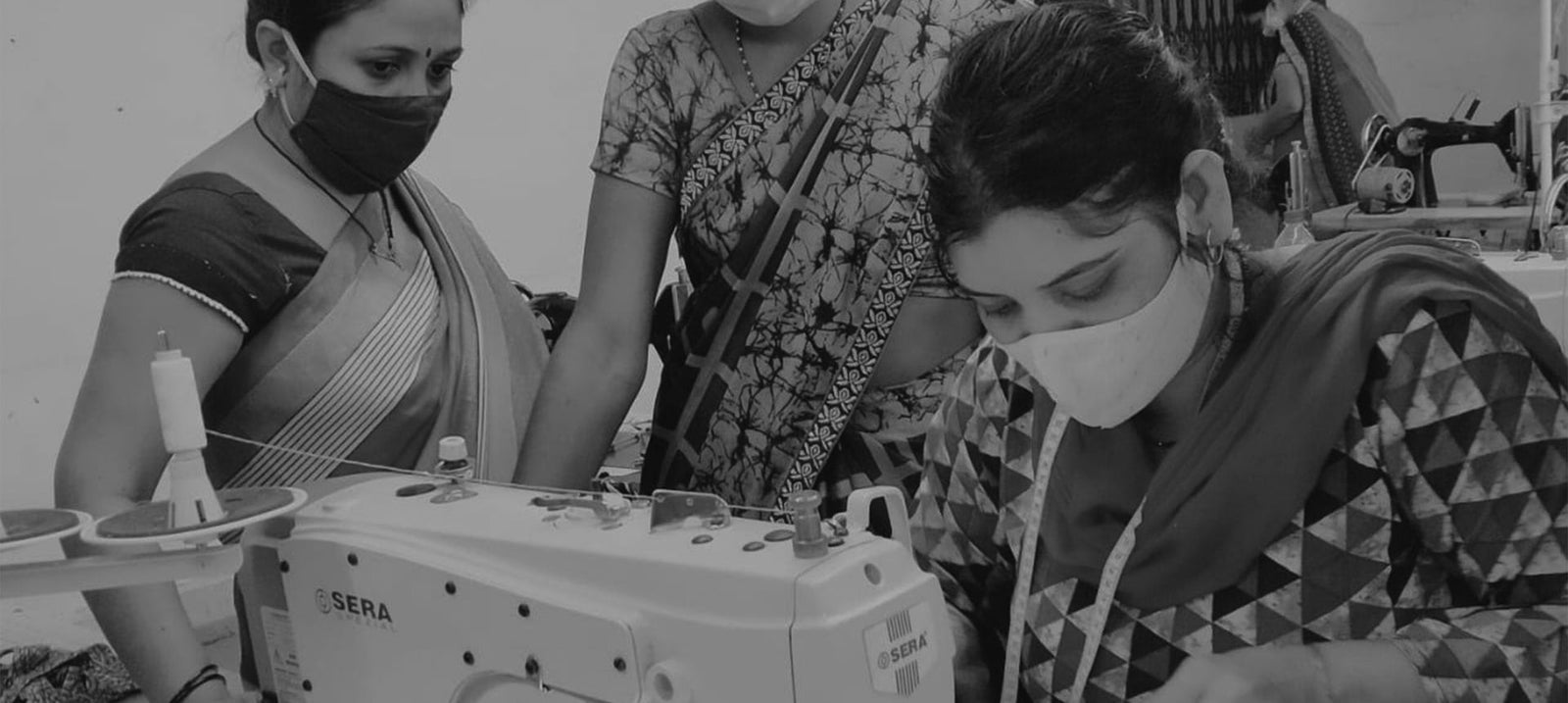 Three women working with sewing machines, wearing masks, in a workshop setting.