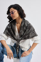 The Mara Scarf in Black styled over a white shirt and blue jeans, worn by a model with pink sunglasses in a casual seated pose.