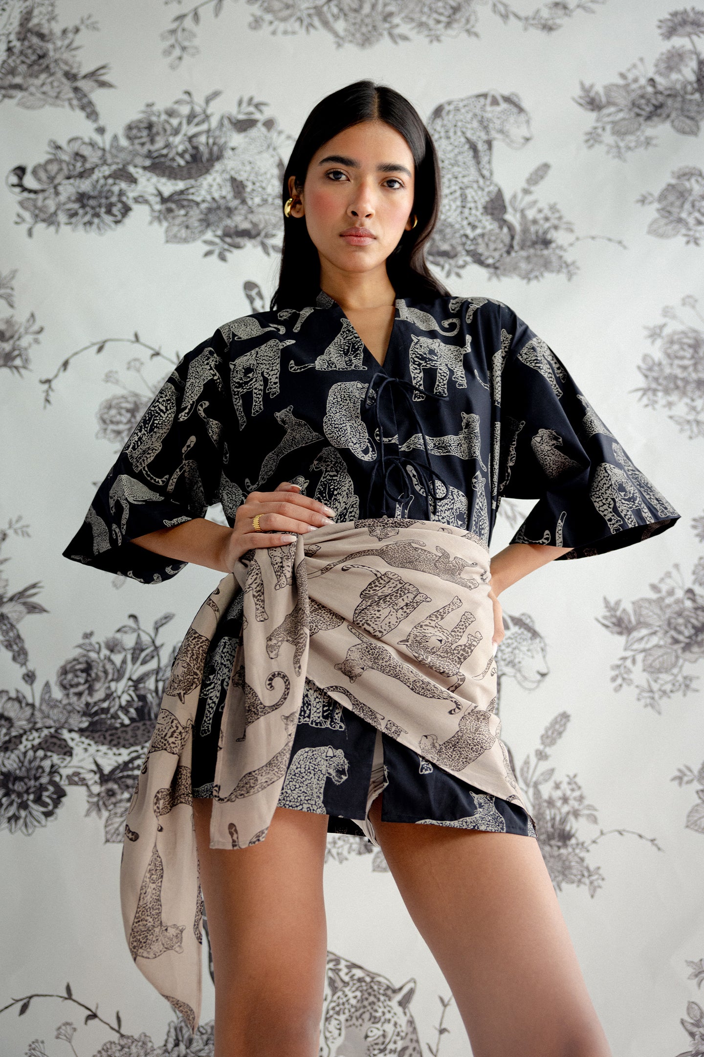 Woman wearing a black and white patterned kimono-style top with a floral background, featuring The Mara Scarf in Brown by Reistor.