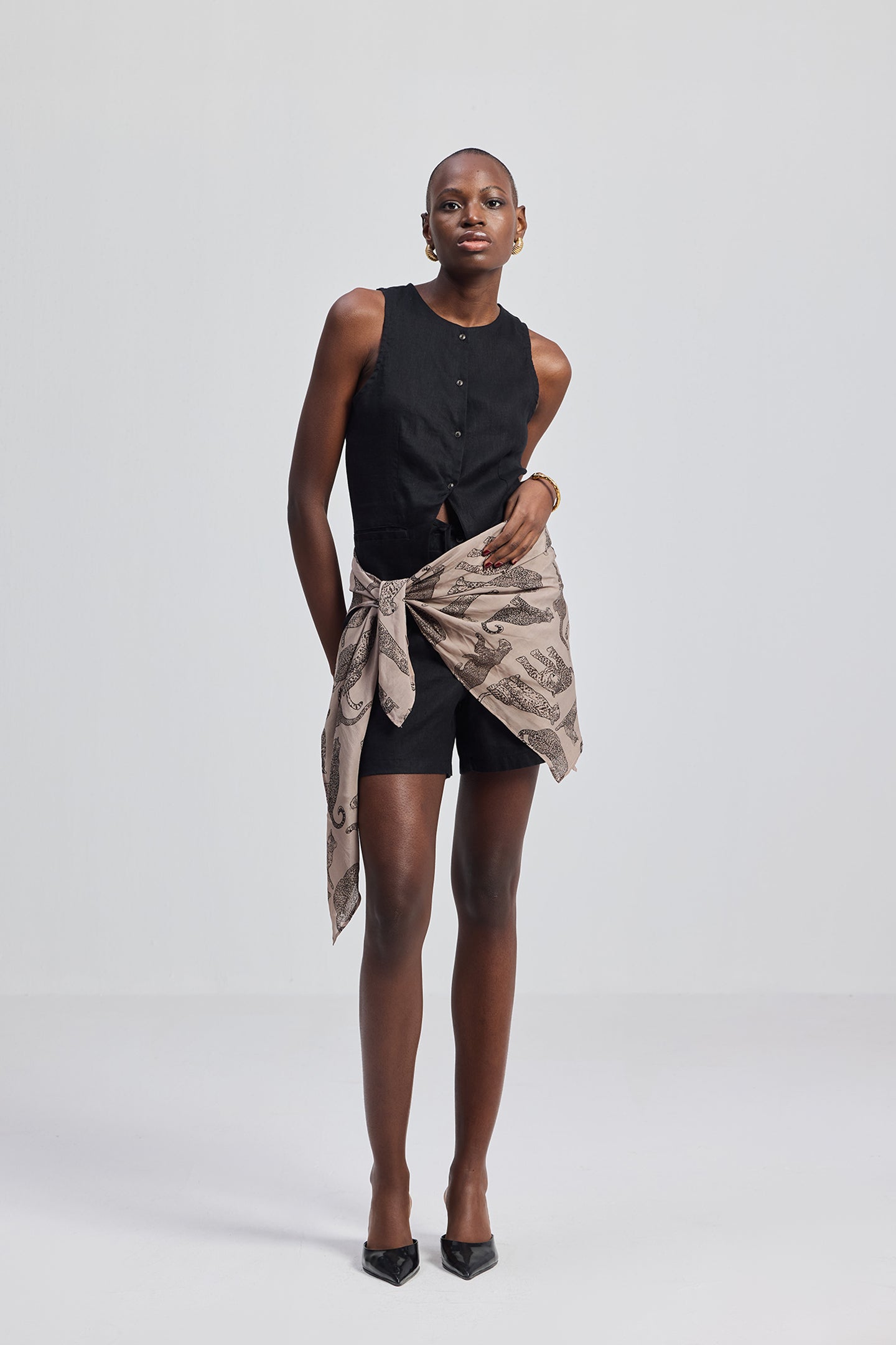 Woman wearing a black sleeveless top and shorts styled with The Mara Scarf in Brown by Reistor on a white background.