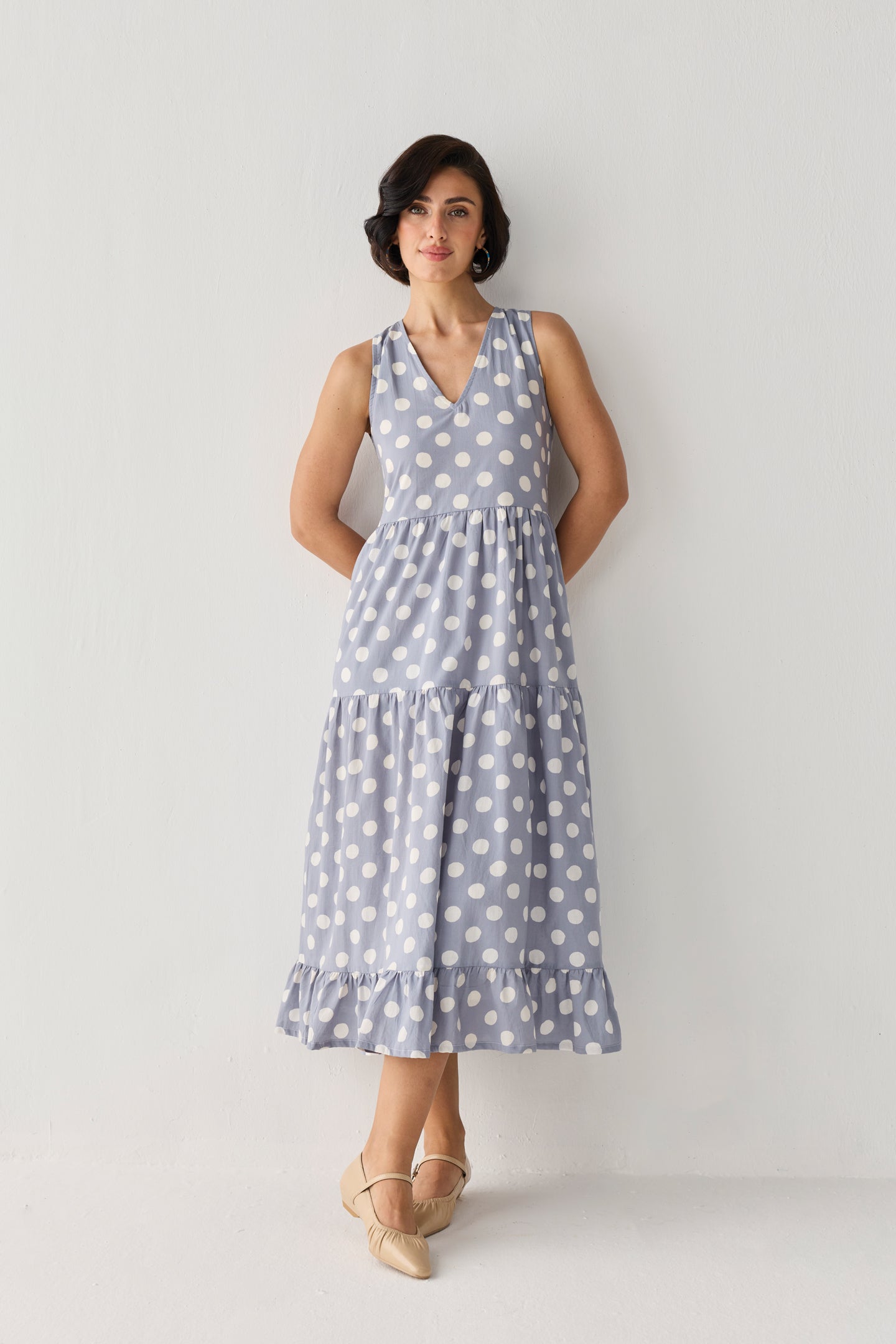 Tiered Midi Dress in Blue Polka with oversized white dots, a sleeveless V-neck bodice, and a ruffled hem, styled in a poised stance with crossed ankles.