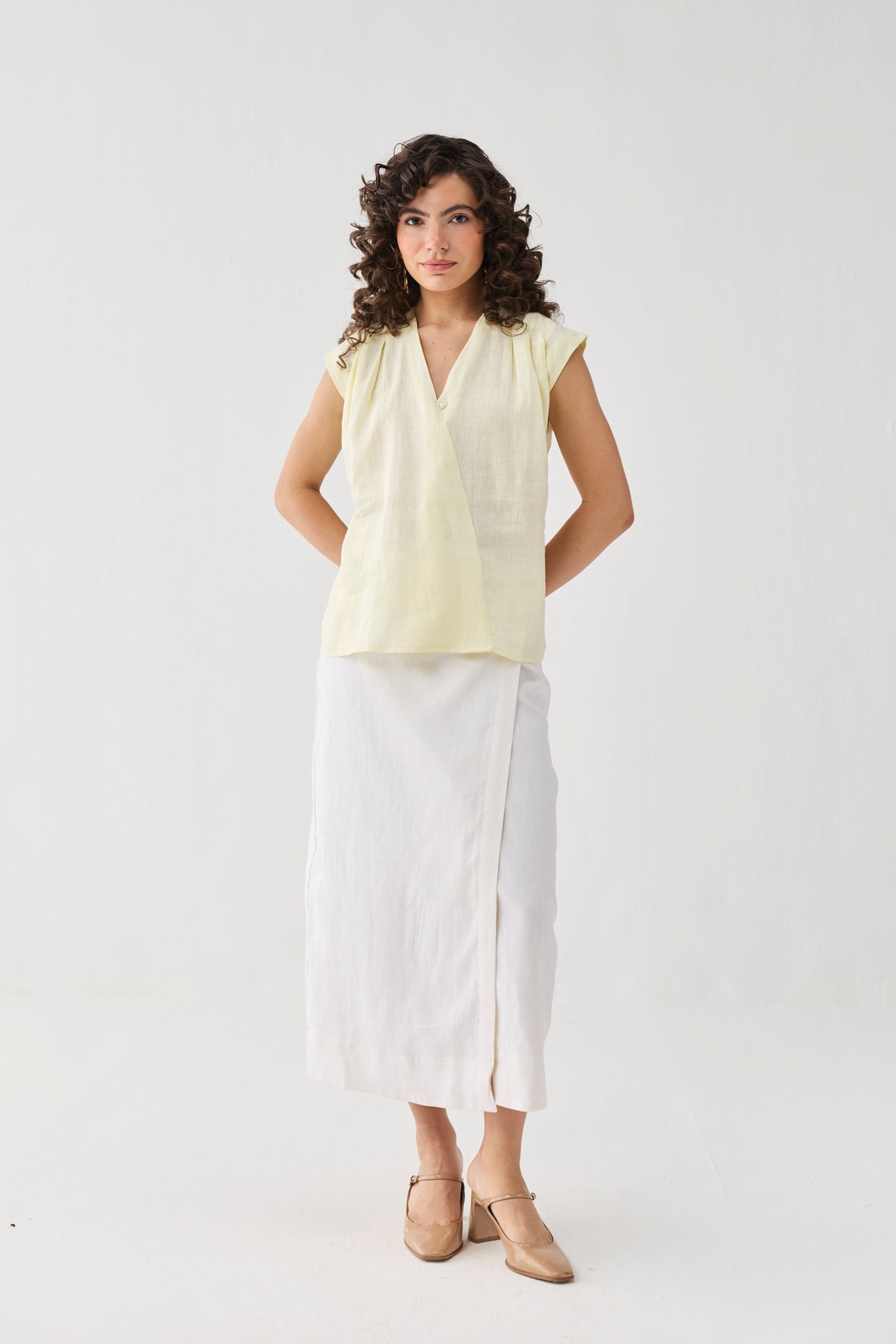 Woman wearing a light yellow sleeveless top and white pants on a white background
