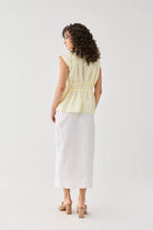 Back view of a woman wearing the V-neck Overlap Top in Butter Lemon with a gathered waist, styled with a white midi skirt and beige block heels. 