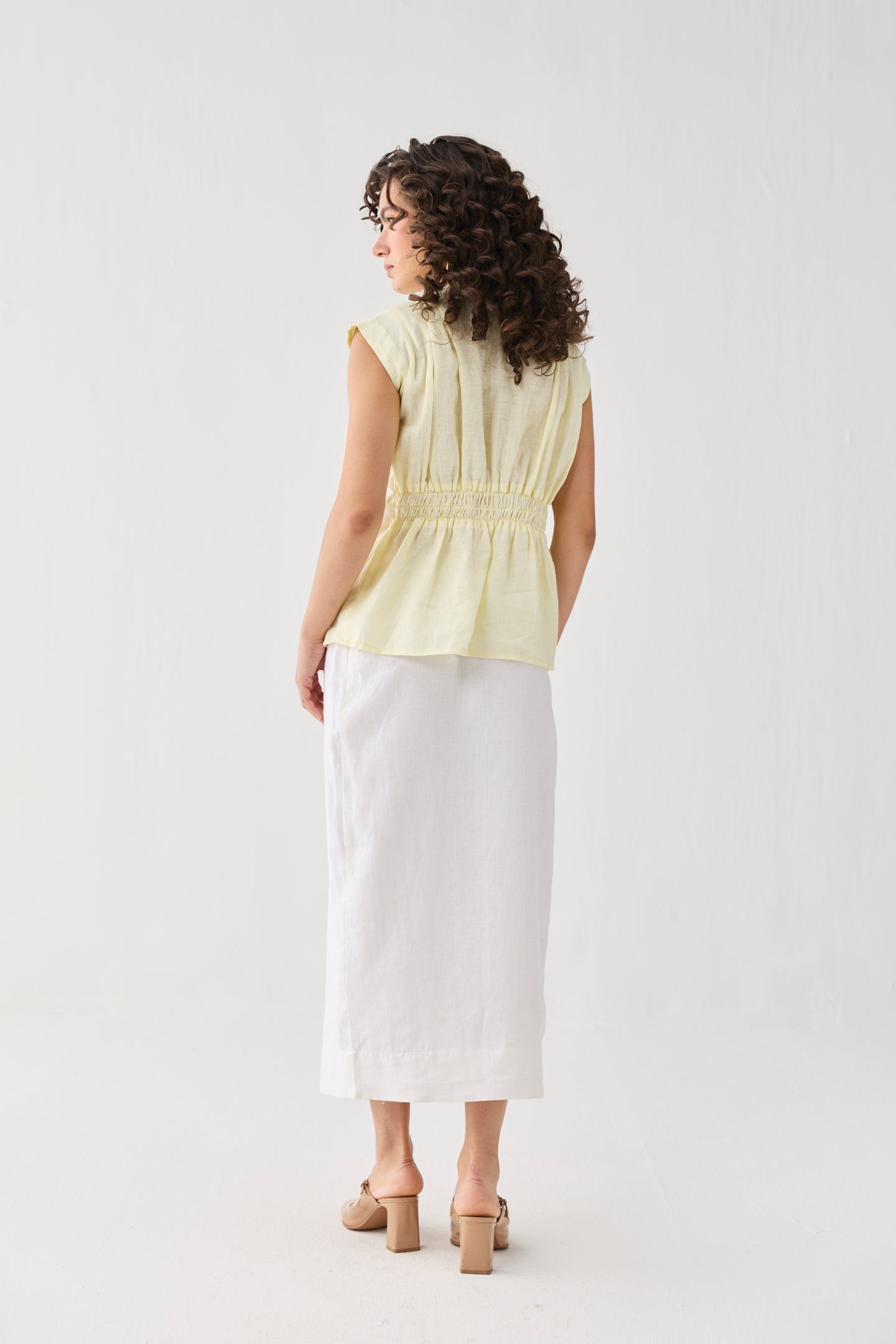 Back view of a woman wearing the V-neck Overlap Top in Butter Lemon with a gathered waist, styled with a white midi skirt and beige block heels. 