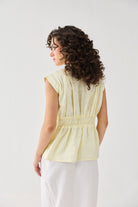 Back view of Reistor's V-neck Overlap Top in Butter Lemon, featuring a smocked waist detail and cap sleeves, styled with a white skirt.