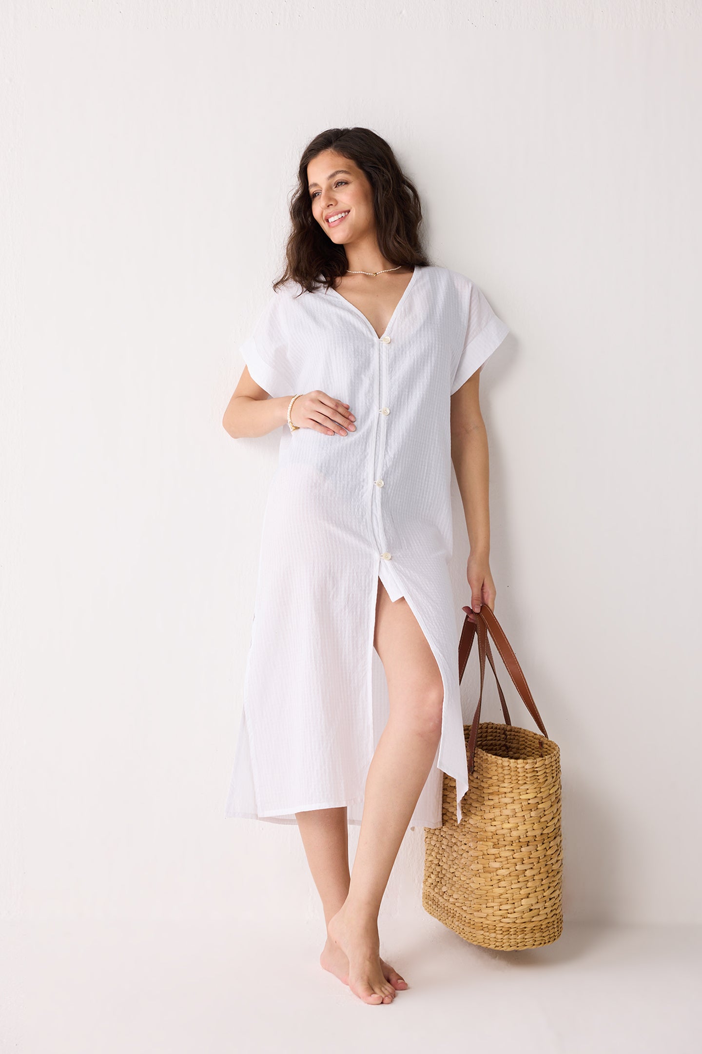 Woman wearing a white cover-up dress holding a straw bag against a white background