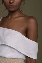 Woman wearing a Reistor white off-shoulder top with gold hoop earrings on a brown background.