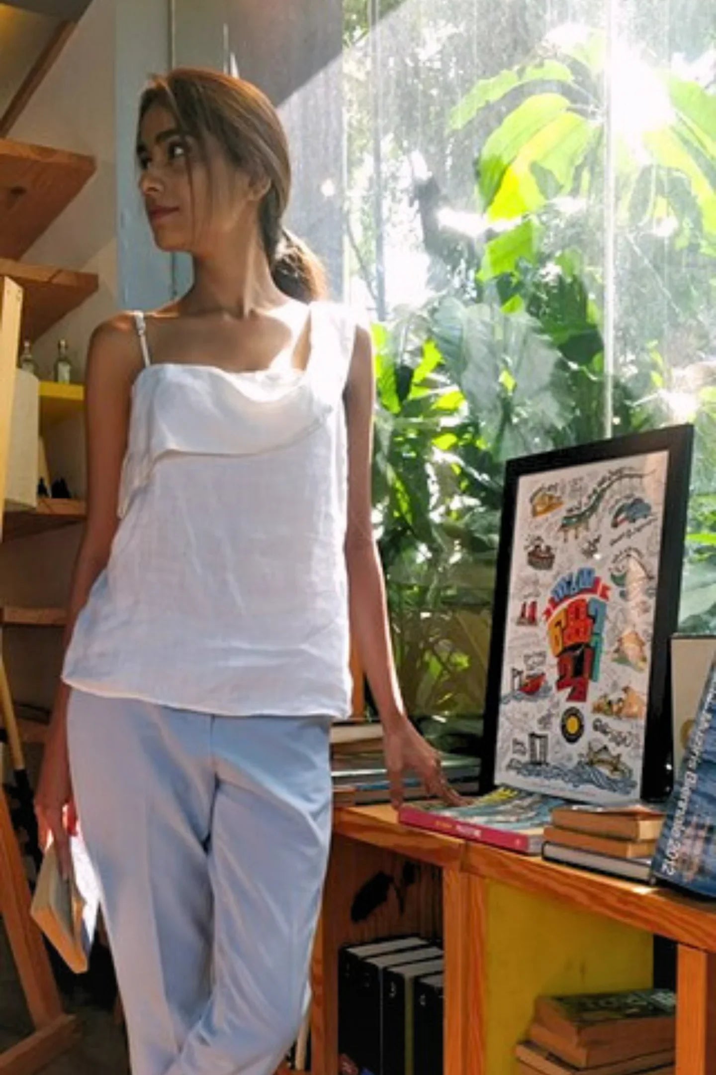 Woman wearing a Reistor white top and light blue pants, standing in a room with books and a framed picture.